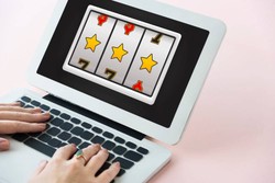 A person types on a laptop showing a slot machine game on the screen, displaying three yellow stars in a row against a soft pink background.