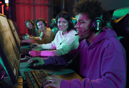 Four young people wearing headsets sit at computers, playing video games together in a dimly lit gaming room. They appear engaged and are smiling, enjoying the group gaming experience.