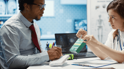 A doctor explains medication instructions to a patient, holding a green and yellow box. The patient listens attentively, sitting at a desk with medical papers, a laptop, and a clipboard.