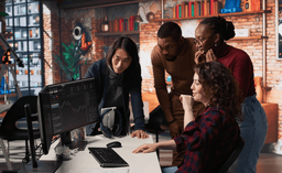 Four colleagues gather around a desk, focused on a computer monitor displaying stock charts and data, in a modern, creative office with brick walls and colorful decor.