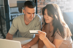 A man and woman sit together at a table, looking at a laptop. The woman holds a credit card while the man looks at the screen, suggesting they are making an online purchase or payment.