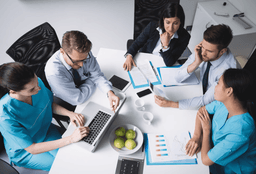 Five medical professionals, some in scrubs and some in business attire, sit around a table with laptops, documents, charts, coffee cups, and a bowl of green apples, engaged in a discussion or meeting.