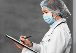 A healthcare professional wearing a lab coat, surgical mask, and hair cover writes on a clipboard, viewed from the side against a neutral background.