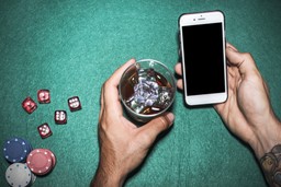 A person holds a glass of whiskey with ice in one hand and a smartphone with a blank screen in the other. Poker chips and dice are on a green felt table.
