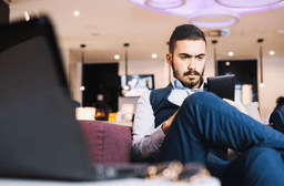 A man with a beard sits on a sofa in a modern lounge, wearing a vest and dress shirt, looking intently at a tablet. A blurred laptop and eyeglasses are visible in the foreground.