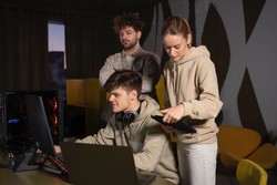 Three young adults in casual clothes work together at a computer desk. One sits with headphones around his neck using a laptop, while the other two stand nearby, looking at a tablet and the screen.