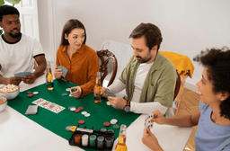 Four people sit around a table playing poker with cards, poker chips, and drinks. They appear engaged in conversation, with snacks and drinks on the table in a brightly lit room.