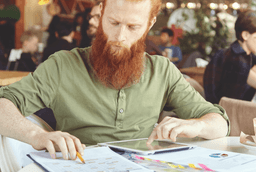 A man with a red beard in a green shirt sits at a table, looking at documents and using a tablet. Papers with charts and a pencil are on the table in a bright, busy indoor setting.