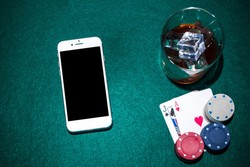 A smartphone, a glass of whiskey with ice, two playing cards (ace and jack of spades), and poker chips are arranged on a green felt surface.