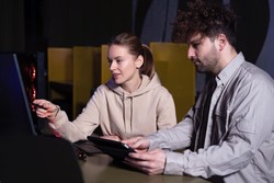 Two people sitting at a desk in front of a large computer monitor; one woman is pointing at the screen while the man beside her holds a tablet. They appear to be discussing something on the computer.