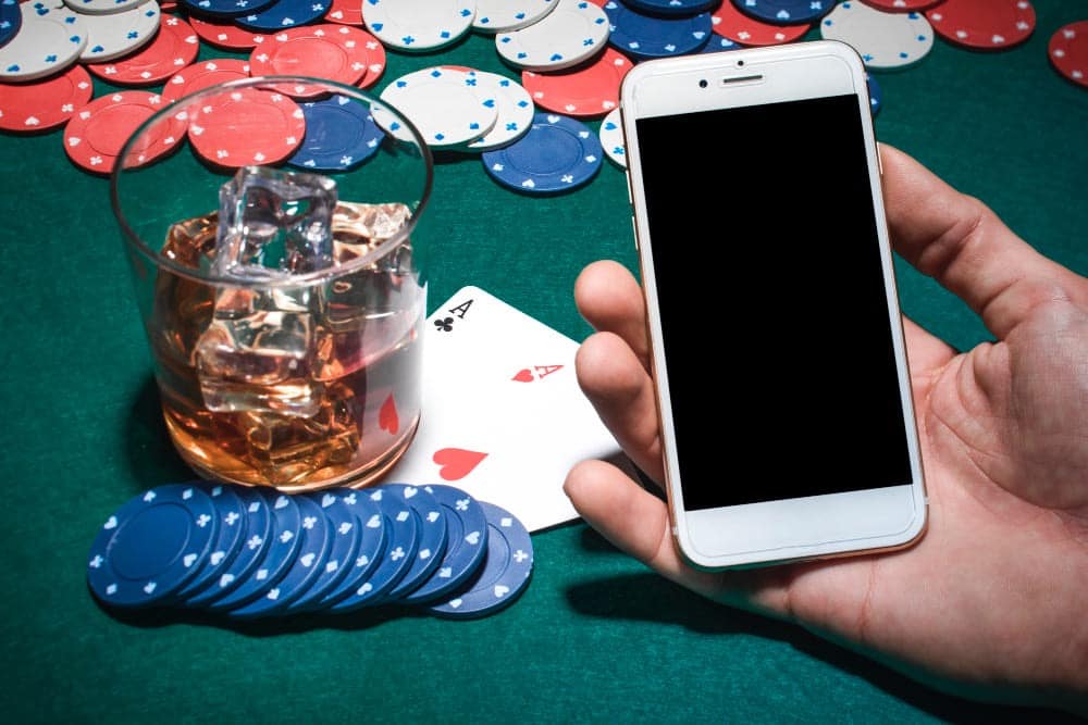 A hand holding a smartphone over a poker table with poker chips, a glass of whiskey with ice, and two playing cards—an ace of spades and ace of hearts—visible on the green felt surface.