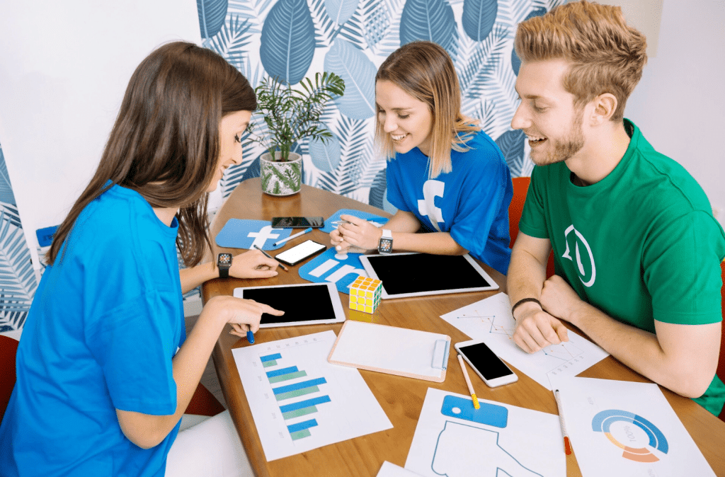 Three young adults sit at a table with charts, smartphones, a tablet, and social media logos, discussing work and smiling. The setting is bright, with patterned wallpaper and a plant in the background.
