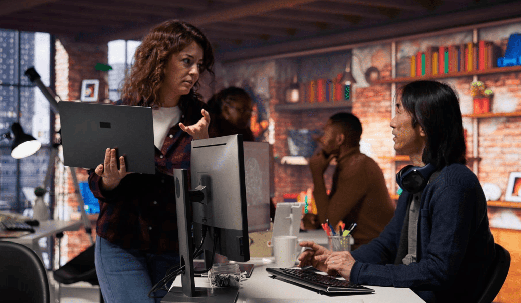 A woman holding a laptop gestures while talking to a man sitting at a desk with a computer monitor in a modern, brick-walled office. Other coworkers are visible in the background.