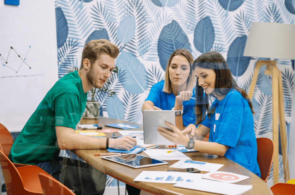 Three young adults sit at a table covered with charts, a tablet, and a laptop, collaborating in a modern office with blue leaf-patterned wallpaper. Two women and one man are focused on discussion and digital devices.